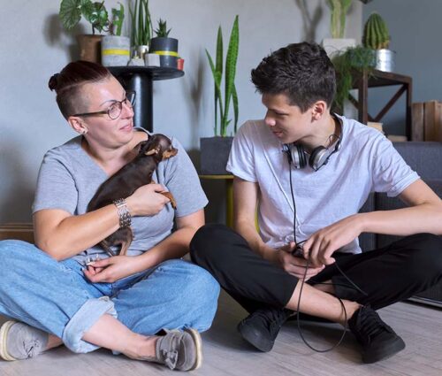 Happy talking mom and teenage son, parent and child sitting at home on the floor Happy talking mom and teenage son, parent and child sitting at home on the floor with pet little dog toy terrier, real friendly loving family